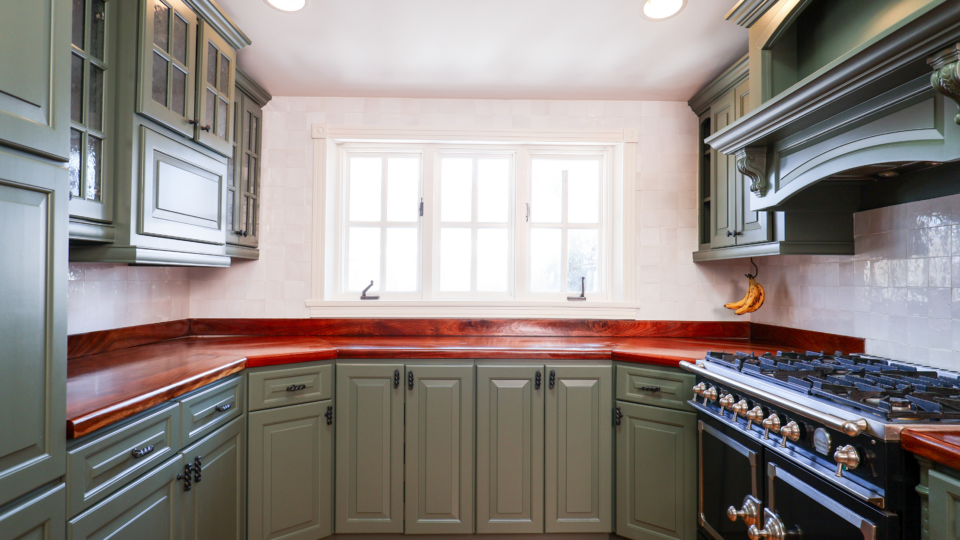 Green cabinetry surrounds a stainless steel pro-style gas range with double ovens and wooden countertops, in a bright kitchen. Banana hangs on the wall nearby.