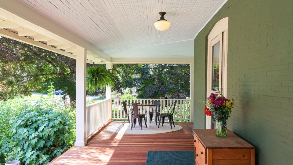 Covered porch with white beadboard ceiling, green brick wall, and a round table with chairs; a vase of flowers on a wooden chest nearby.