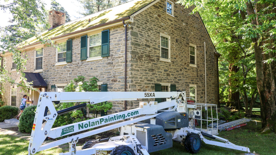 Stone two-story house with green shutters being serviced by a painting crew; a large white boom lift labeled Nolan Painting is in the yard, trees surrounding the scene.