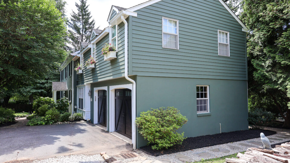 Green two-story house with white trim, black garage doors, and flower planters beneath windows.