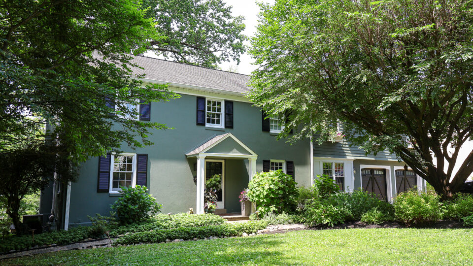 Two-story green house partially hidden by dense trees in a lush front yard from the street view.