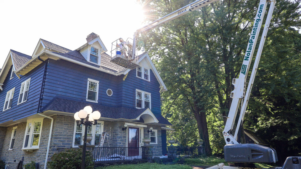 Worker in a bucket lift painting the upper roofline of a blue house beside trees and a lawn.