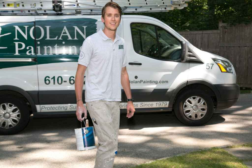 Painter stands in front of a white van with green Nolan Painting branding and a ladder on top.