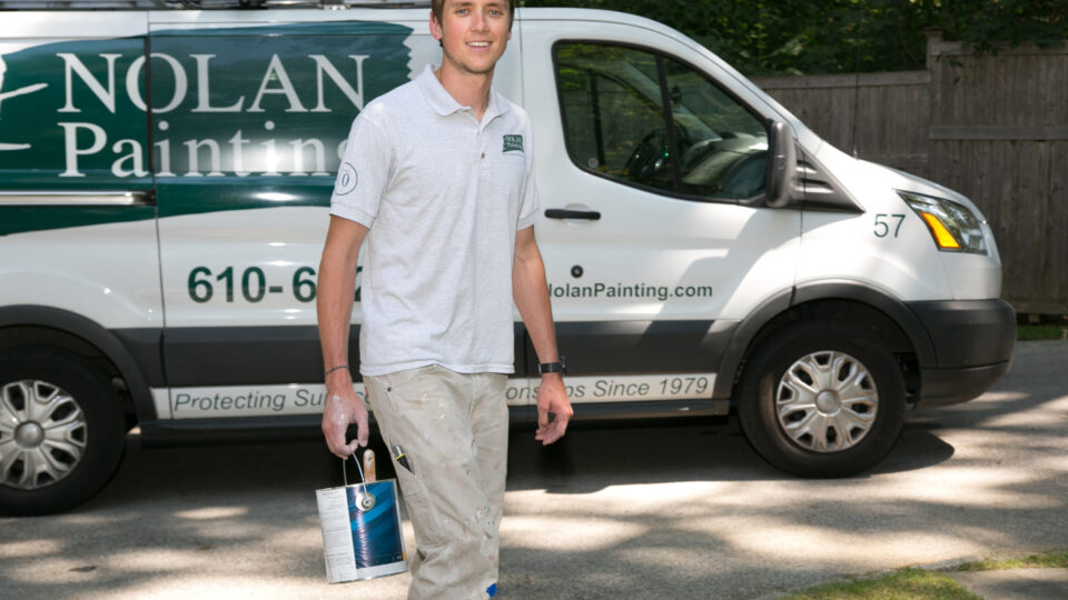 Painter stands in front of a white van with green Nolan Painting branding and a ladder on top.